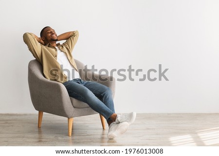 Weekend. Happy African American Man Relaxing Sitting In Chair Holding Hands Behind Head Posing With Eyes Closed On Gray Wall Background, Enjoying Silence And Comfort Of Minimalism Lifestyle Royalty-Free Stock Photo #1976013008