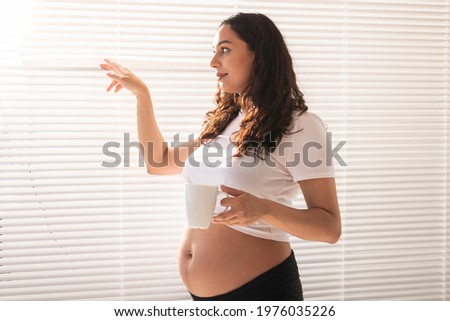 Curious young beautiful pregnant woman drinking tea and looking through the blinds at the window. Concept of joy and good news while waiting for baby