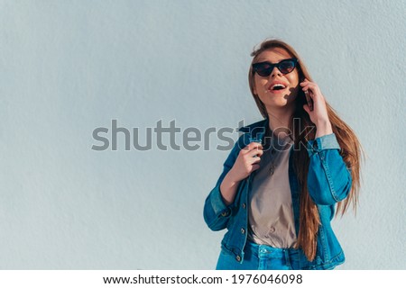 Young beautiful woman using a smartphone and wearing jeans and sunglasses with a white wall in the background