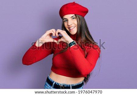 Young brunette teenager wearing french look with beret smiling in love doing heart symbol shape with hands. romantic concept. 