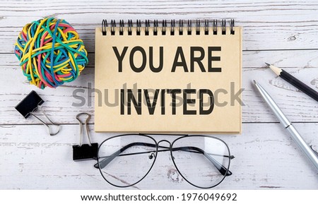 Notebook with text YOU ARE INVITED and office supplies. View from above. Closeup on the white background