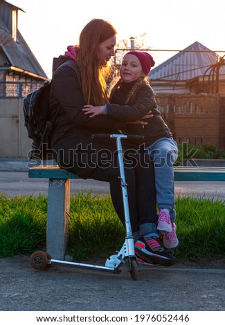 Mother and daughter on a bench in the park. Child riding on kickscooter. Blonde cute girl on scooter spring city street background. Active lifestyle sports leisure. New normal lockdown social distance