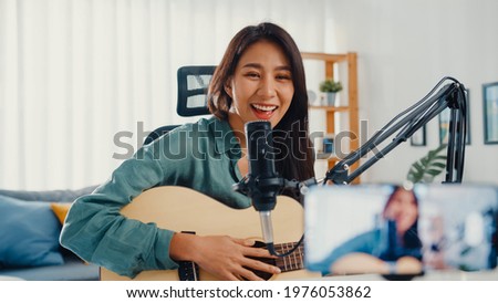 Teenage Asia girl influencer play guitar music use microphone record with smartphone for online audience listen at home. Female podcaster make audio podcast from her home studio, Stay at home concept. Royalty-Free Stock Photo #1976053862