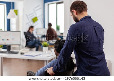 Businessman helping his disabled black colleague entering in company office pushing wheelchair near desk. African handicapped paralysed woman greetting colleagues starting working at financial project