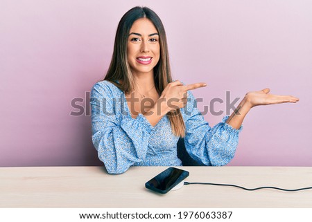 Young hispanic woman wearing casual clothes charging smartphone battery amazed and smiling to the camera while presenting with hand and pointing with finger. 