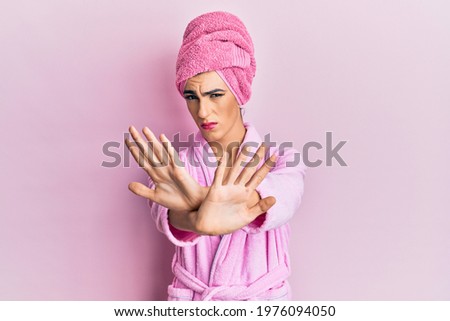Young man wearing woman make up wearing shower towel on head and bathrobe rejection expression crossing arms and palms doing negative sign, angry face 
