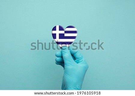 Hands wearing protective surgical gloves holding Greece flag heart