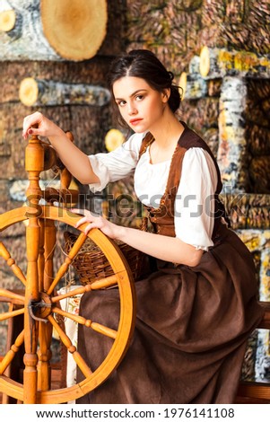 Young Lovely Caucasian Brunette Woman Posing With Spinning Wheel in Retro Dress In Village Environment. Vertical Shot