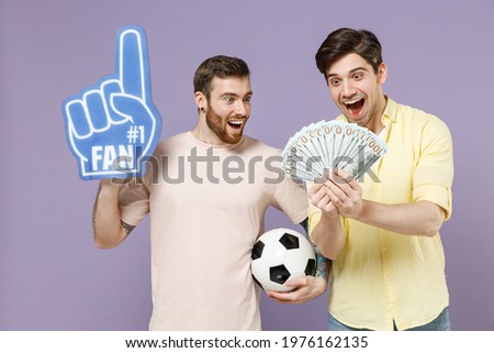 Two young shocked men friends together in casual t-shirt fan foam glove finger hold fan cash money in dollar banknotes soccer football ball isolated on purple background. People lifestyle concept.