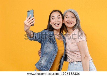 Excited cheerful family asian female women girls gray-haired mother brunette daughter in casual clothes posing doing selfie shot on mobile phone isolated on yellow color background studio portrait