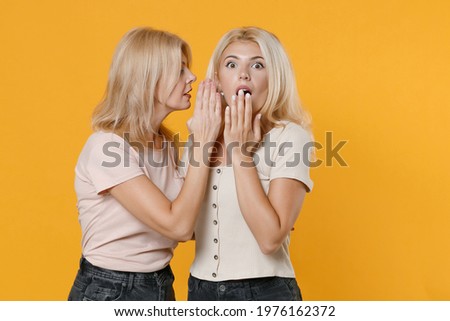Shocked family two women blonde mother daughter in casual clothes hugging whispers gossip and tells secret with hand gesture covering mouth isolated on bright yellow colour background studio portrait
