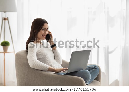 Covid-19 pandemic, new normal and work remote. Happy friendly millennial attractive arabian woman with laptop speaks on phone, sits in armchair, on window background, in living room interior, profile