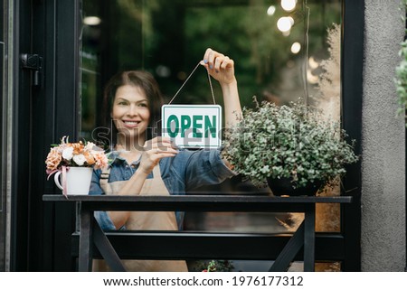 End of quarantine. Welcoming guests after coronavirus outbreak. Happy young pretty european female in apron turning open sign board on glass door in modern flower studio, eco cafe or restaurant