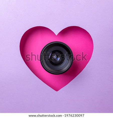 Pink heart on a purple background. At the heart is a camera lens. Concept of love, photography and video.