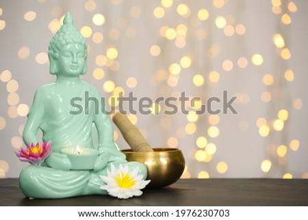 Buddha statue with candle, lotus flowers and singing bowl on table against blurred lights. Space for text