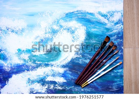 Painter paint acrylic on canvas with brush with blue ocean on frame. Artwork as hobby or occupation for artist. Big wave in sea w creates artwork by mix color and decors picture as hard copy art.