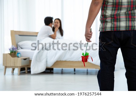 Asian husband in scottish shirt holding heart branch seeing wife and male adulterer on bed in bedroom Royalty-Free Stock Photo #1976257544