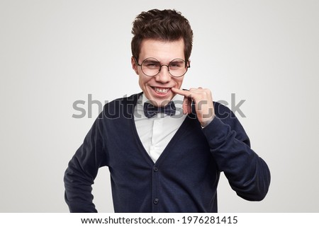 Young gentleman in classic outfit with bow tie and nerdy spectacles making funny face while smiling and looking at camera against white background