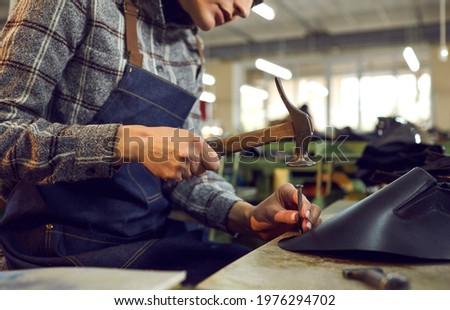 Shoe factory worker using professional tools. Cropped shot woman sitting at workshop table using hand hammer and nail to make new leather boots. Footwear production and manufacturing industry concept