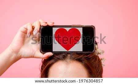 Take a picture of a young woman putting a heart-shaped smartphone on her head. Love concept