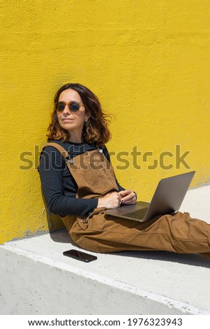 Lifestyle woman with sunglasses sitting on the floor and leaning against the wall on the street on a sunny day working with laptop and phone.