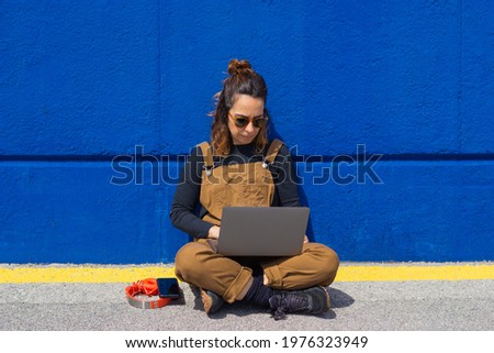 Lifestyle woman with sunglasses sitting on the ground and leaning against the wall on the street on a sunny day taking a selfie and working with laptop and headphones by her side.