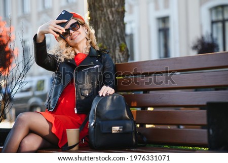 Cheerful young woman takes selfie, sits on wooden bench outside and takes selfie in good mood.
