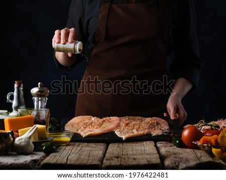 Turkey steak. The cook salts the pieces of meat. Cooking process. Delicious nutritious Thanksgiving meal. On the table are tomatoes, lemon, salt, pepper. Dark background. Close-up. Royalty-Free Stock Photo #1976422481