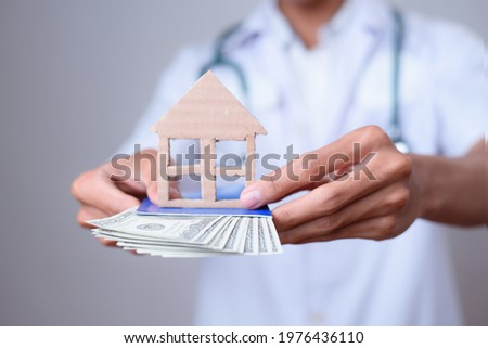 Close up of male doctor hands showing money, banknotes and shapes of miniature house isolated gray background. Tax and insurance concept