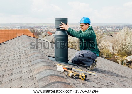 worker with blue helmet on the roof installing iron chimney. roofing Construction and Building New iron House with Modular Chimney. Royalty-Free Stock Photo #1976437244