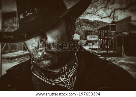 Old West Cowboy Western Town. An old west cowboy in a hat smoking a hand rolled cigarette with an old western town setting in the background, edited in vintage film style. Royalty-Free Stock Photo #197650394