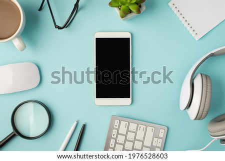 Top view photo of smartphone in the middle cup mouse pens magnifier keyboard headphones notepad plant and glasses on isolated pastel blue background