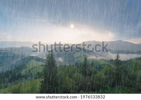 Rain and sun through the clouds over the green summer Carpathian foggy mountain hills. Rainy evening panorama. Royalty-Free Stock Photo #1976533832
