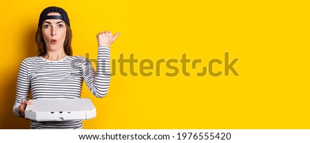 surprised young woman pizza delivery man, points finger to the side while holding pizza on yellow background. Banner.