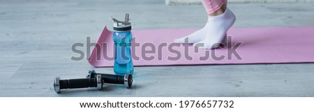 cropped view of woman in socks on fitness mat near dumbbells and sports bottle, banner