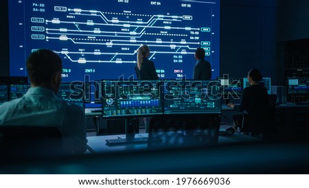 Project Manager and Computer Science Engineer Talking while Using Big Screen Display Showing Infrastructure Infographics and Data.Telecommunications Company System Control and Monitoring Room Royalty-Free Stock Photo #1976669036