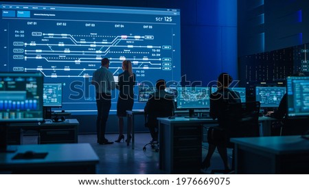 Telecommunications Company System Control and Monitoring Room with Diverse Multicultural Team of Professionals Working on Computers. Big Screen Display Showing Infrastructure Infographics. Back View Royalty-Free Stock Photo #1976669075