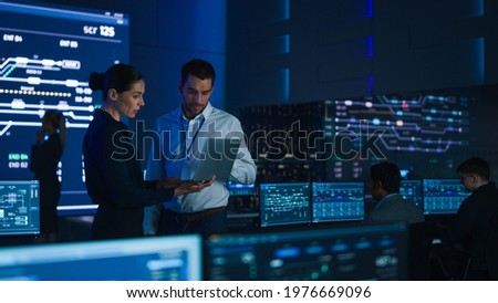 Project Leader and Chief Executive Discuss Data Shown on a Laptop. In Background Big Screen Showing Infographics of Infrastructure, Charts, System Analysis. Telecommunications Control Room Royalty-Free Stock Photo #1976669096