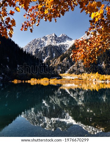 Mountain landscape of autumn forest with yellow trees and snowy white peak with reflection on Lake Issyk in Kazakhstan