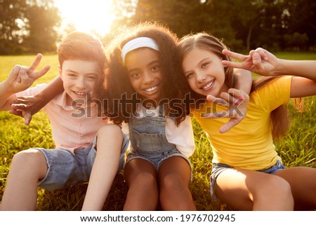 Company of happy diverse children sitting on grass and hugging while showing two fingers gesture and looking at camera