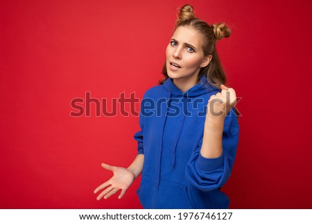 Photo of emotional agressive young beautiful blonde woman with two horns with sincere emotions wearing hipster bright blue hoodie isolated over red background with free space and showing fist