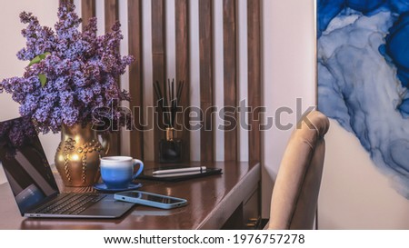 A cup of tea or coffee on a working wooden table, a laptop, a phone, a bouquet of lilacs in a gold vase. Cozy interior with a blue accent. Working office desk, working at home. 