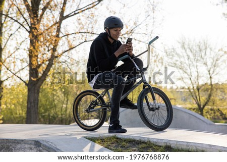 Dark-skinned teenager sits on bmx bike in park, skatepark propping himself up with one leg and texts with friends via social media, waits for mates