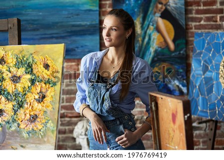 Portrait of young smiling female artist. Painter with her artworks in art studio. Creative process, relaxation, leisure, hobby, stress management