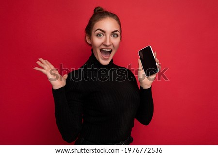 Photo of attractive overjoyed positive young brunette woman wearing black sweater standing isolated over red background showing mobile phone with empty screen for mockup looking at camera