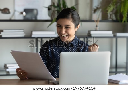 Overjoyed millennial Indian female employee triumph read good news work online on laptop in office. Happy young ethnic woman worker feel euphoric with pleasant hire notice or message in paper letter. Royalty-Free Stock Photo #1976774114
