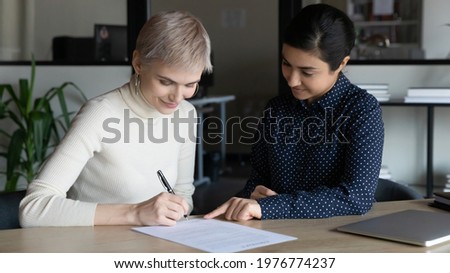Happy young Caucasian woman sign paper document close deal with Indian business partner or client at meeting. Smiling multiracial businesswomen put signature on paperwork contract make agreement.