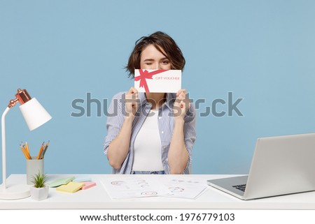 Young fun successful employee business woman in casual shirt sit work at white office desk with pc laptop hiding cobver mouth with gift voucher flyer mock up isolated on pastel blue background studio