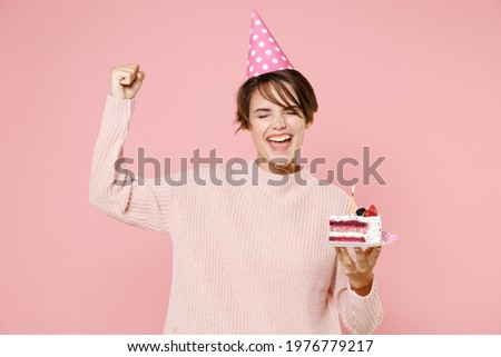 Happy fun young brunette woman 20s in knitted casual sweater birthday hat isolated on pastel pink background. Birthday holiday party people emotions concept. Celebrating hold cake doing winner gesture