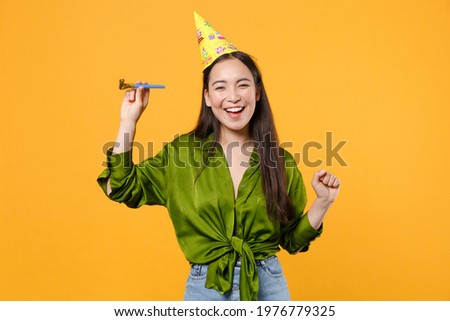 Funny cheerful beautiful young brunette asian woman wearing basic green shirt birthday hat hold pipe dancing clenching fist looking camera isolated on bright yellow colour background, studio portrait
